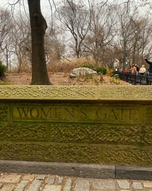 A low stone wall at the border of central park. Trees and an autumn leaf-strewn ground are behind it. To the side is a black fence and benches and a few people enjoying the crisp day in the park.