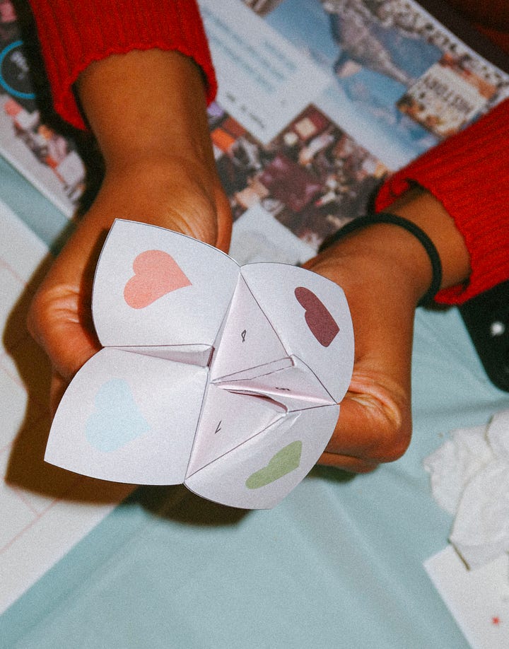 Hands holding a paper fortune teller decorated with pastel hearts; the fortune teller laid out against a green background.
