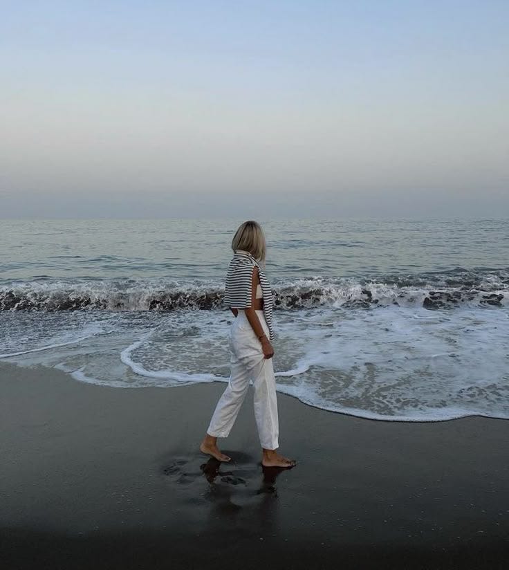 This may contain: a woman is walking along the beach with her feet in the water and looking at the ocean This may contain: a woman is walking along the beach with her feet in the water and looking at the ocean