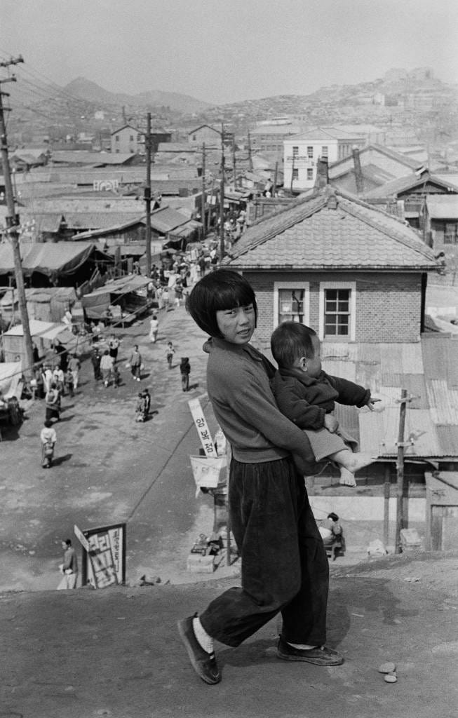 Black-and-white photo of a Korean girl carrying a baby on her hip, walking along a dirt ridge above a dense neighborhood in 1950s Seoul. Below her, narrow streets are crowded with market stalls, low tin and tile roofs, utility poles, and people moving through the postwar city.