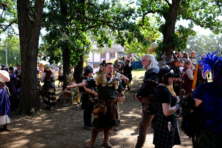 Bards singing in a tavern wearing medieval garb, an elf and wizard surveying goods at a shop, fire-twirler show, Geralt and another warrior deep in conversation, a man playing dead with an arrow under his armpit, guests walking toward the camera, a woman with a horse head looking over a table.