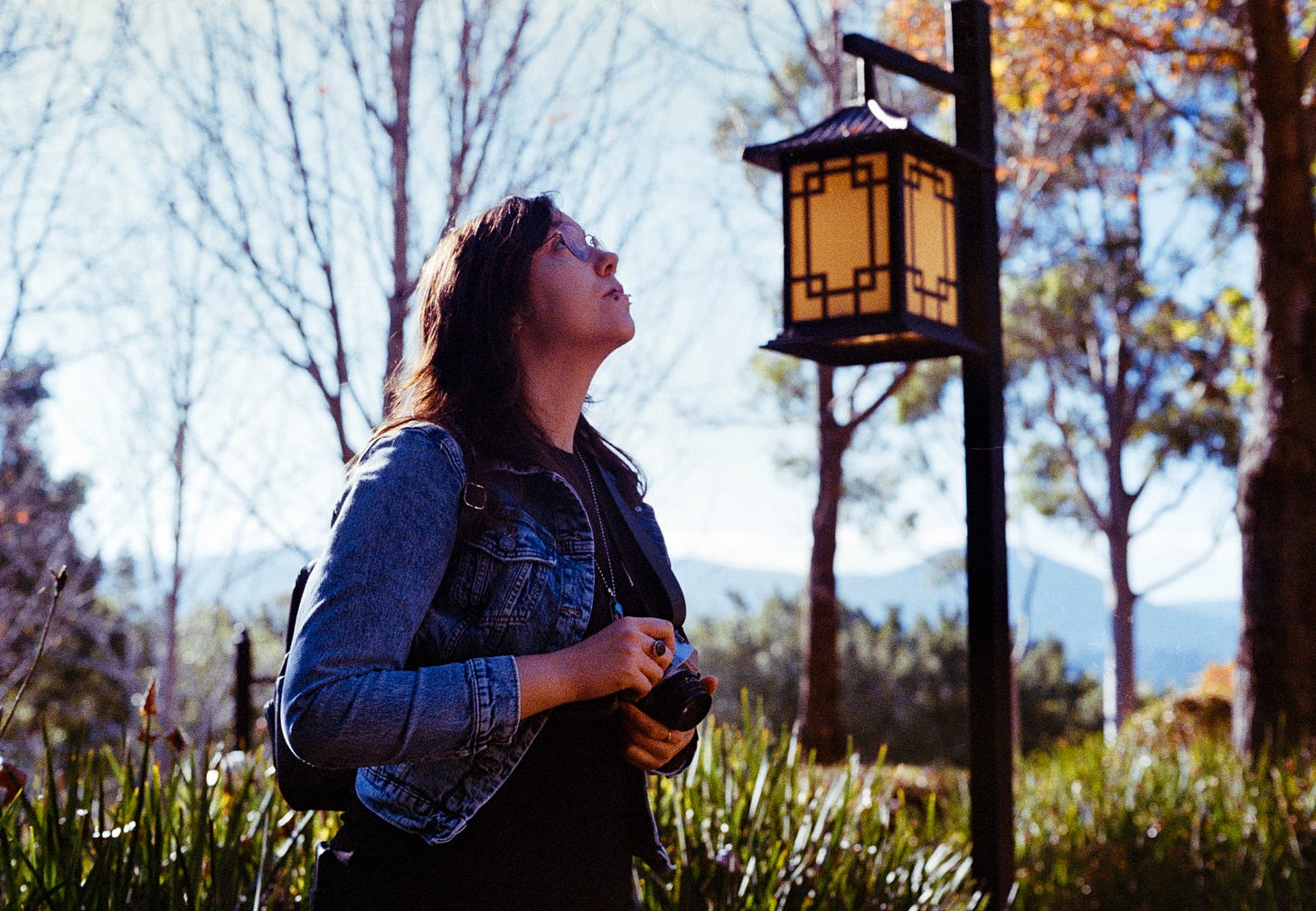 A photo of Josie, wearing her signature denim jacket, holding a Pentax camera and looking up, contemplating a new angle, a different shot. A photo of Josie, wearing her signature denim jacket, holding a Pentax camera and looking up, contemplating a new angle, a different shot.