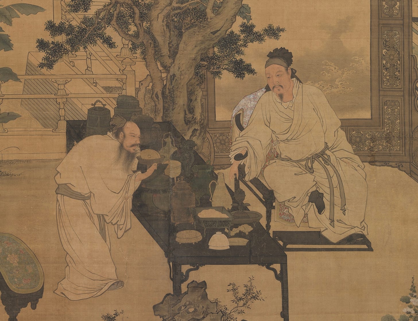 A section of the Ming Dynasty painter Tu Chin’s scroll painting, “Enjoying Antiquities.” The painting shows two gentlemen in an elegant chamber regarding a long, rectangular table of antiquities. One stands, and is removing the lid of a small ceremonial pot. The other is seated on the opposite side of the table. Behind him is a painting in an elaborate frame, and a small, gnarled tree. Objects on the table include vases, bowls, pots and small statues.