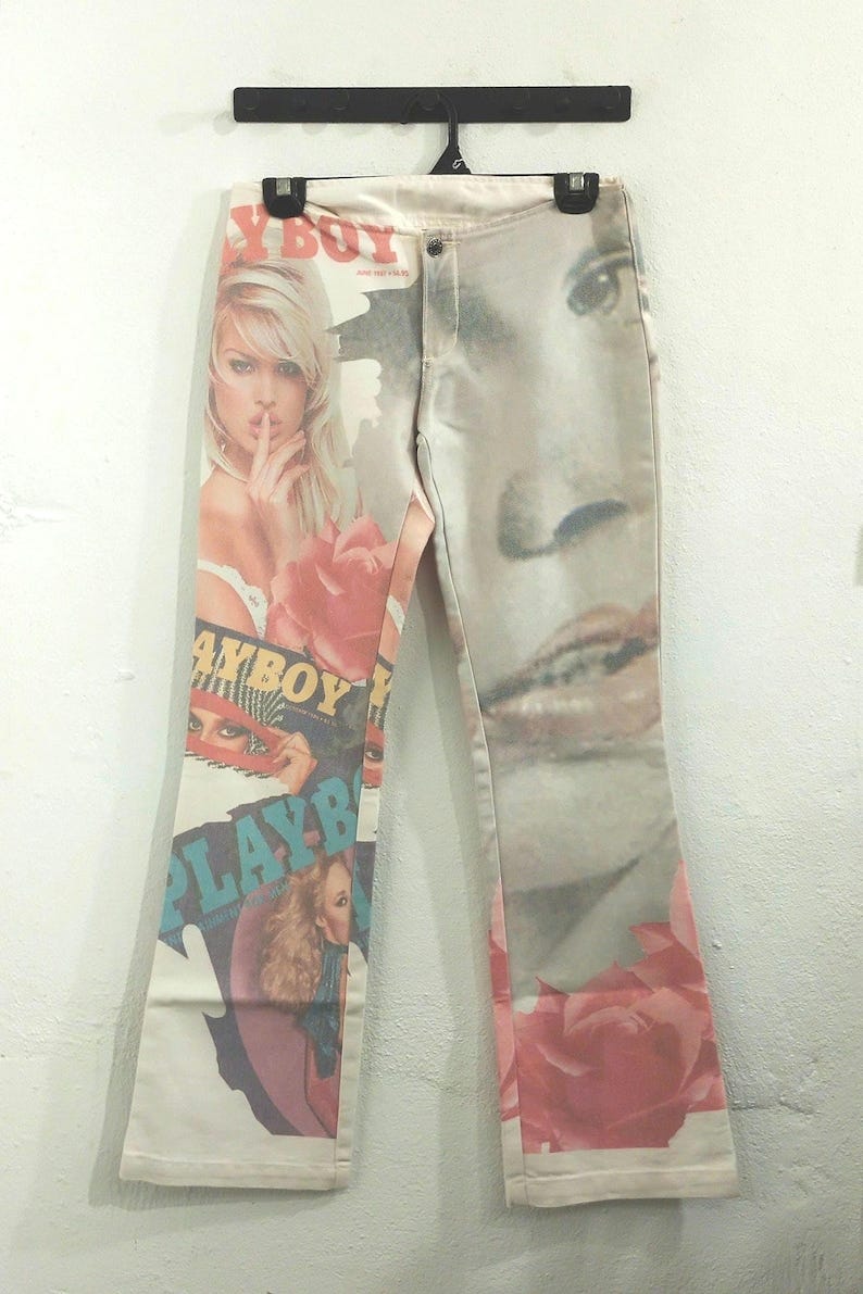 May include: Off-white denim pants with a printed design featuring images of women, roses, and the text "PLAYBOY". The pants are hanging against a white wall. May include: Off-white denim pants with a printed design featuring images of women, roses, and the text "PLAYBOY". The pants are hanging against a white wall.