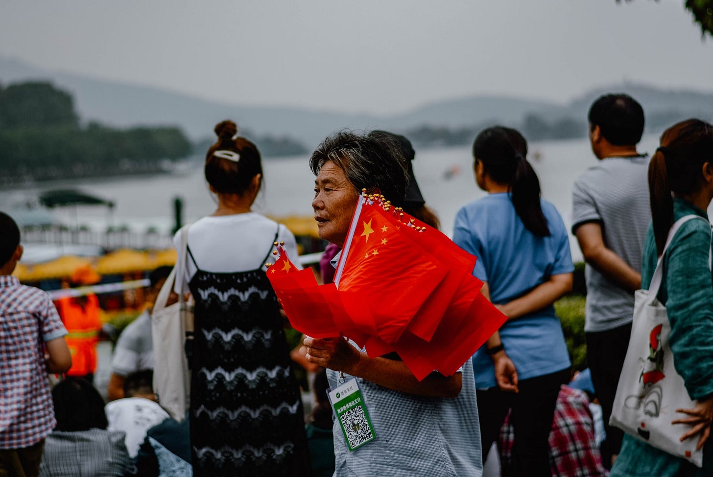 Fotografia em cores de idoso chinês carregando bandeiras da China. Ao fundo, pessoas observam um rio.