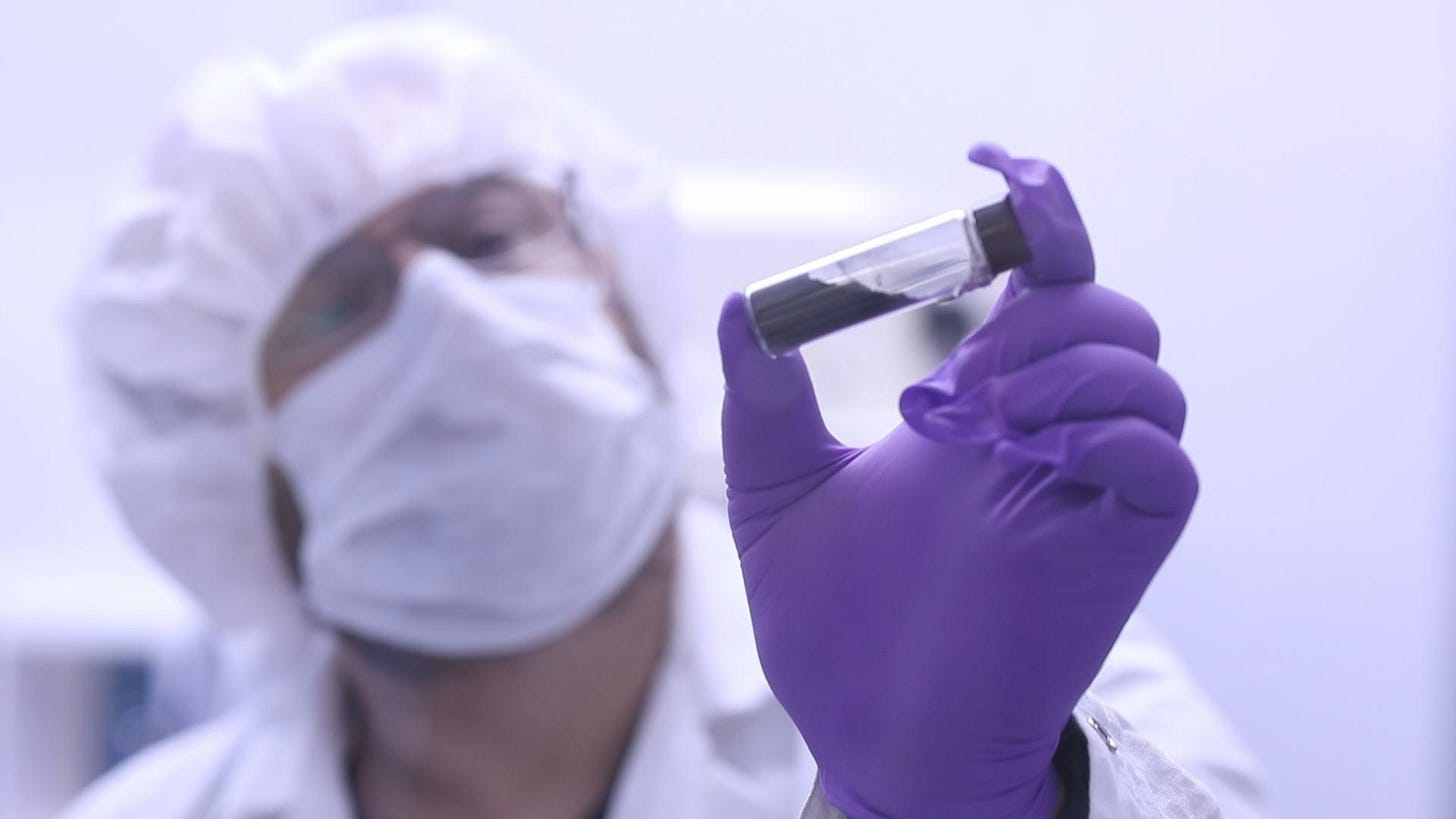 A scientist wearing protective clothing and purple gloves holds up a test tube for examination in a laboratory setting. A scientist wearing protective clothing and purple gloves holds up a test tube for examination in a laboratory setting.