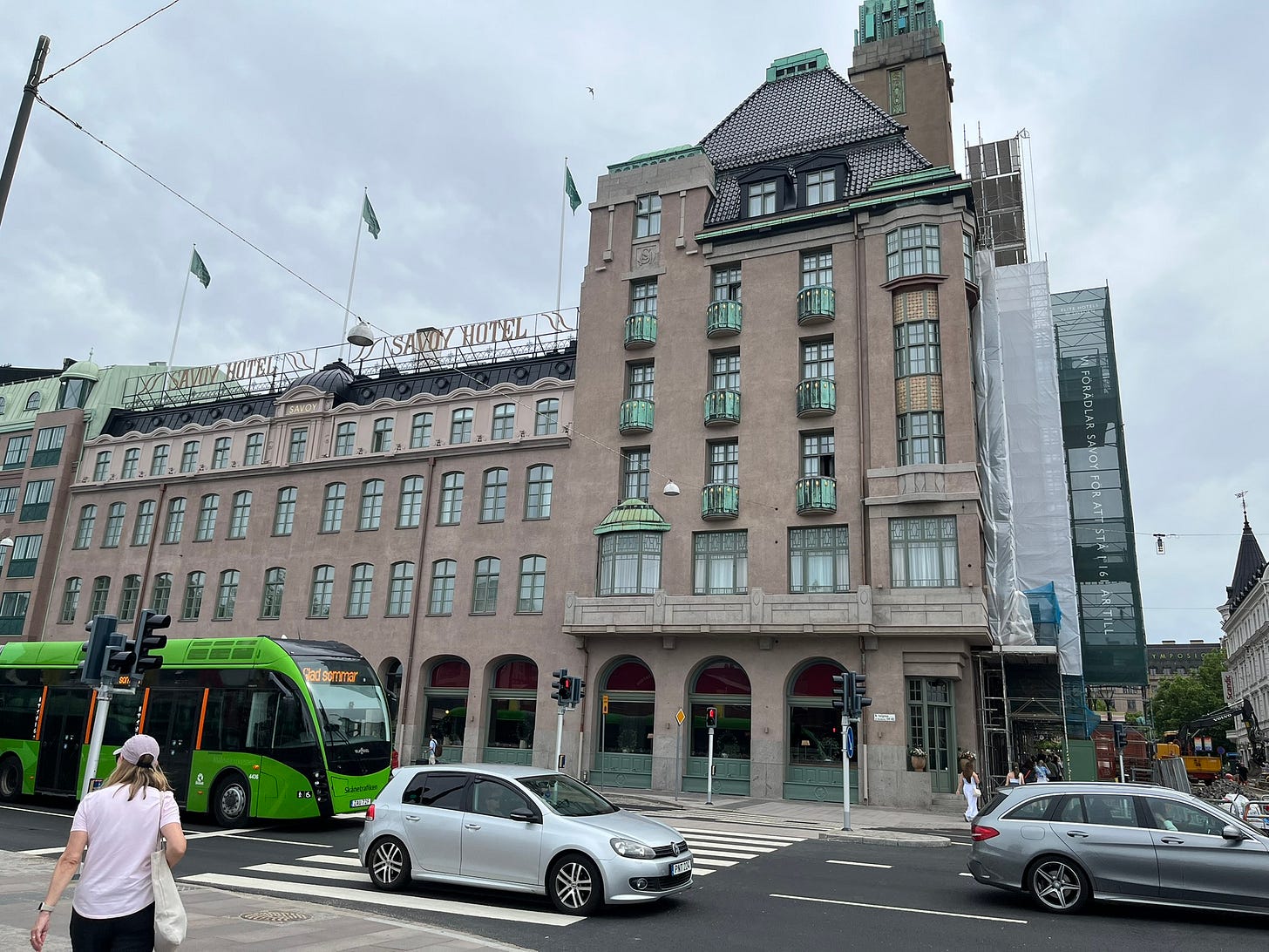 A wide light brown building with 4 stories to the left, and 6 stories to the right. There is a dark gray triangular roof to the right, with a short, thin green tower behind it. In the middle of the building, there is a golden sign saying "SAVOY HOTEL" on top. In front of the building is a road with 2 cars and a green bus.