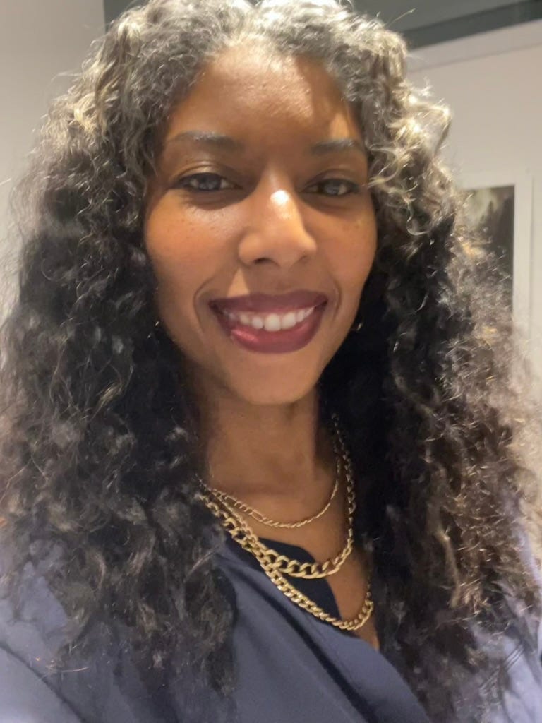 Khadijah wearing a dark blue silk v-neck top, smiling. Her hair is very long and curly, with about three inches of white and gray at the roots, with the rest still black. She is wearing gold jewelry and dark red lipstick.