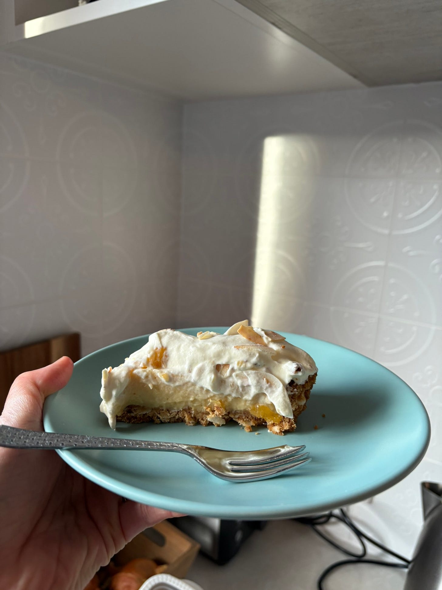 A slice of mango cream pie, topped with toasted coconut. It's on a pale blue plate, held up in a kitchen with sun hitting the plate