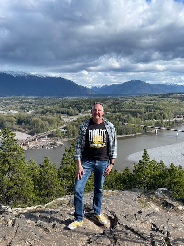 Dr. Dave in flannel on top of a hill with beautiful mountains and a cloudy sky behind him