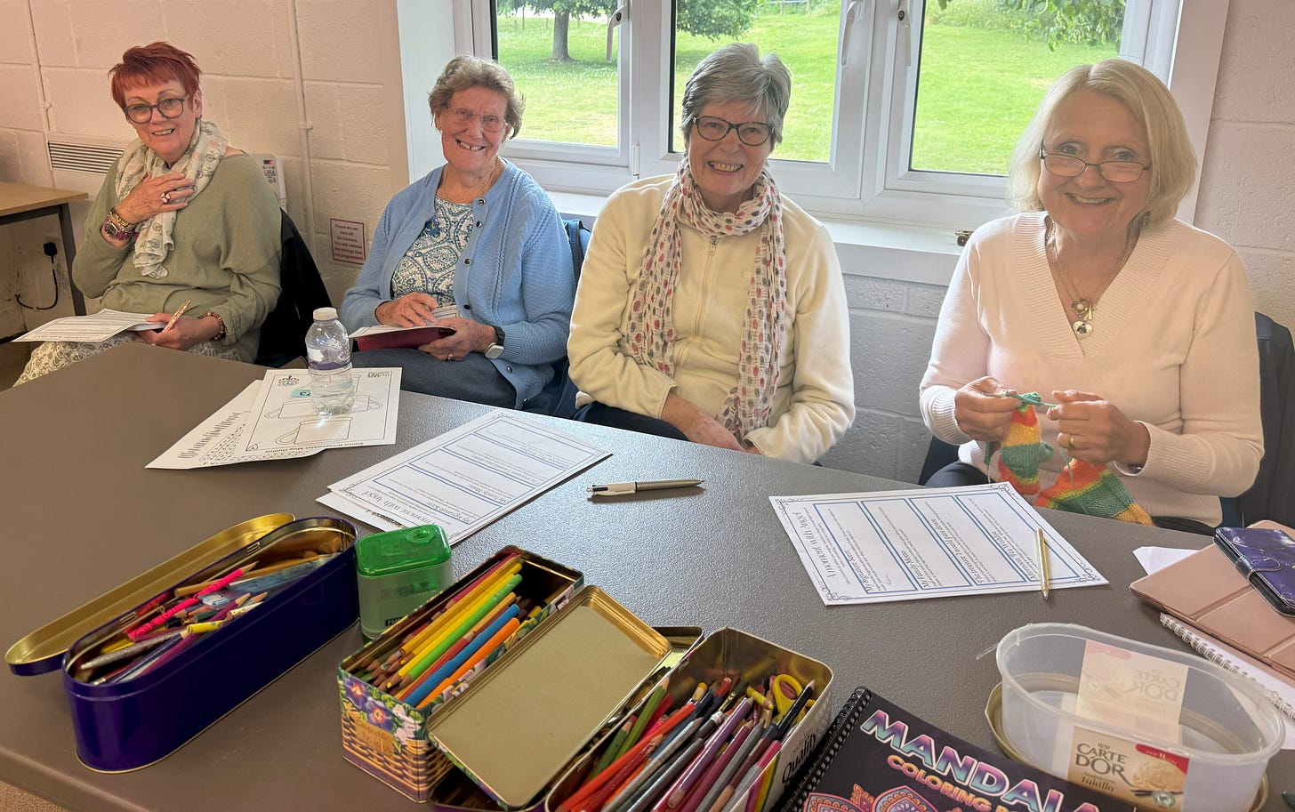 Four women are seated at a table, smiling in a bright room. In front of them are coloring supplies, paper, and a knitting project, creating a cozy, creative atmosphere.