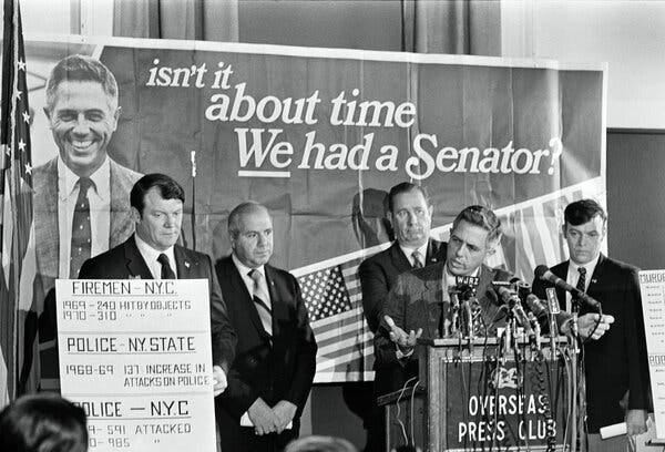 A black-and-white photo of Mr. Buckley, who stands at a podium festooned with microphones and gestures with his with right hand. Four serious-looking men with suits and ties stand behind hin, in front of a large sign with his photo and the words “isn’t it about time We had a Senator?,” with the word “We” underlined. A black-and-white photo of Mr. Buckley, who stands at a podium festooned with microphones and gestures with his with right hand. Four serious-looking men with suits and ties stand behind hin, in front of a large sign with his photo and the words “isn’t it about time We had a Senator?,” with the word “We” underlined.