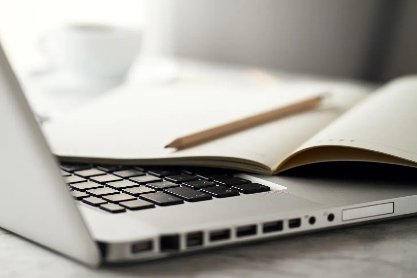 An open laptop computer with a journal and pencil lying on top. Blurred in the background is a tea cup.