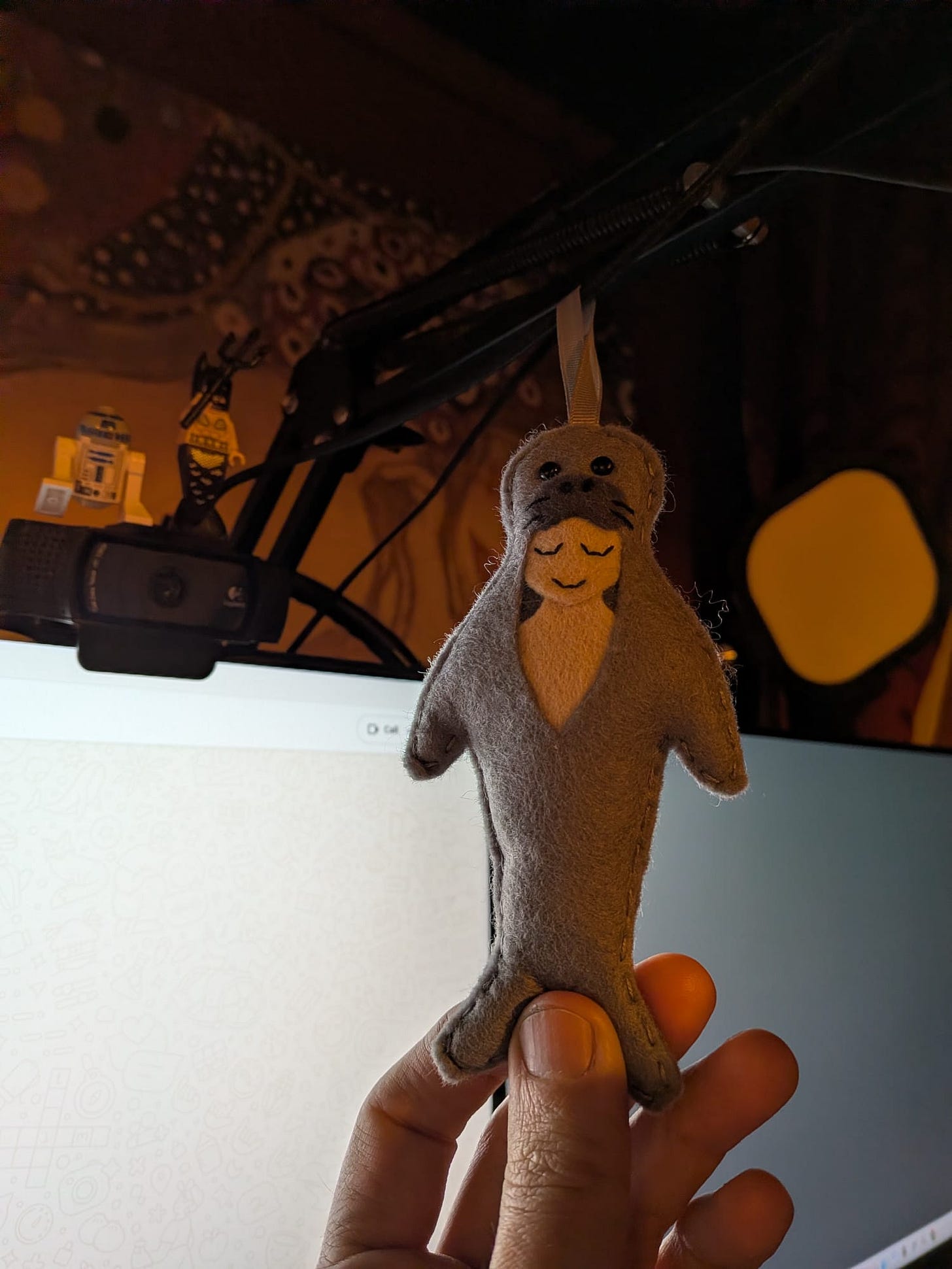 Handmade soft felt sea creature or seal-like doll held in front of a computer setup, photographed as a whimsical personal object during Tarot DMs with Emma van Straaten.