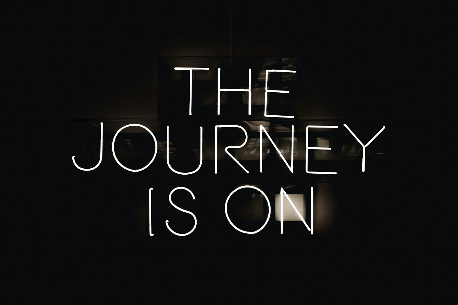A black background with a minimalist neon sign displaying the text "THE JOURNEY IS ON" in white light. The text is arranged in three lines with "THE" on top, "JOURNEY" in the middle, and "IS ON" at the bottom, creating a simple but striking illuminated message against the dark surroundings.
