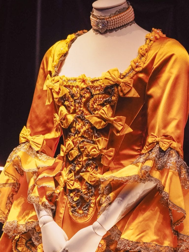 A dazzling yellow period-style dress adorned with delicate bows, worn by Taylor Swift in the Bejeweled music video. The gown features intricate detailing, evoking an elegant historical aesthetic, and is complemented by a pearl necklace, adding a touch of timeless sophistication.