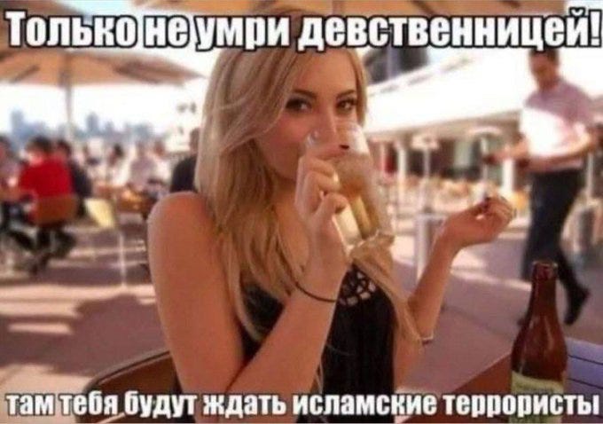 A woman with long blonde hair wearing a black sleeveless top, holding a glass of beer and a bottle of beer, sitting at an outdoor table. People and a cityscape are visible in the background.