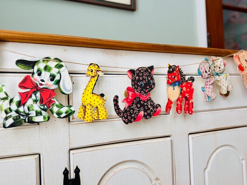 May include: A garland of vintage-style animal cutouts, including a dog, giraffe, cat, and horse, strung on twine. The dog has a green and white checkered pattern, the giraffe is yellow with brown spots, and the cat is black with pink floral details. May include: A garland of vintage-style animal cutouts, including a dog, giraffe, cat, and horse, strung on twine. The dog has a green and white checkered pattern, the giraffe is yellow with brown spots, and the cat is black with pink floral details.