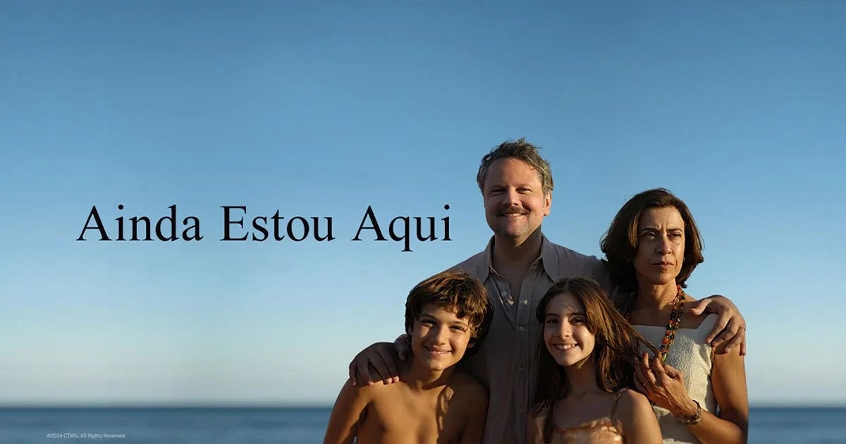 Quatro pessoas sorrindo em frente ao mar, com o céu azul ao fundo. Texto: 'Ainda Estou Aqui'.