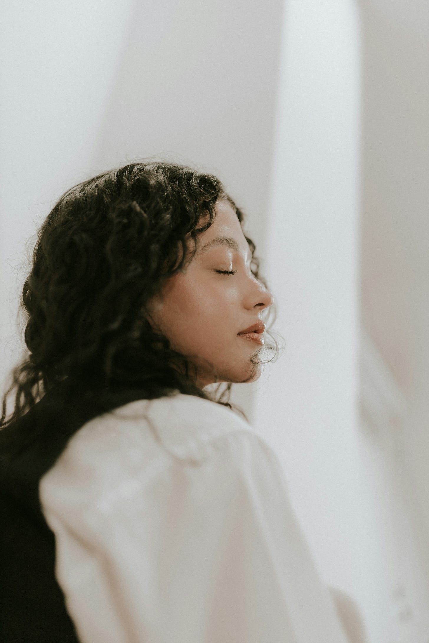 Profile of a woman with curly hair and eyes closed, wearing a white top. The soft light and serene expression create a calm, peaceful mood. Profile of a woman with curly hair and eyes closed, wearing a white top. The soft light and serene expression create a calm, peaceful mood.
