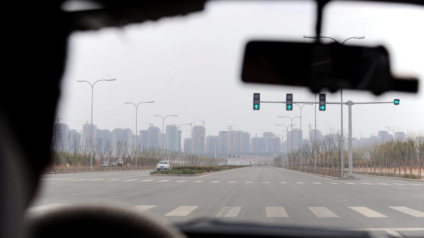View from inside a vehicle looking out at a wide, empty road with green traffic lights and modern skyscrapers in the background, under a cloudy sky.