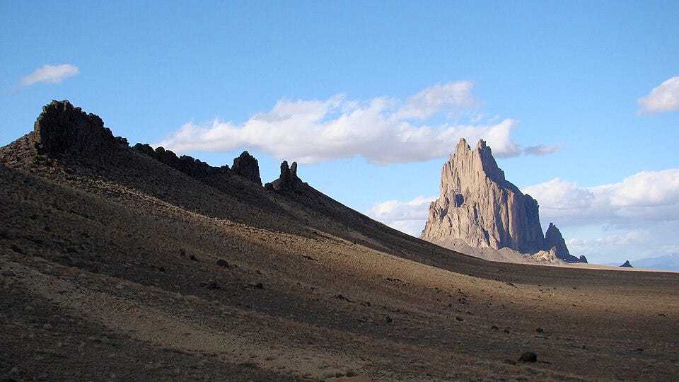 In the background is Shiprock, a giant vertical protrusion of rock. In the foreground is a magmatic dike, a smooth incline from the upper left to the lower right of the screen.