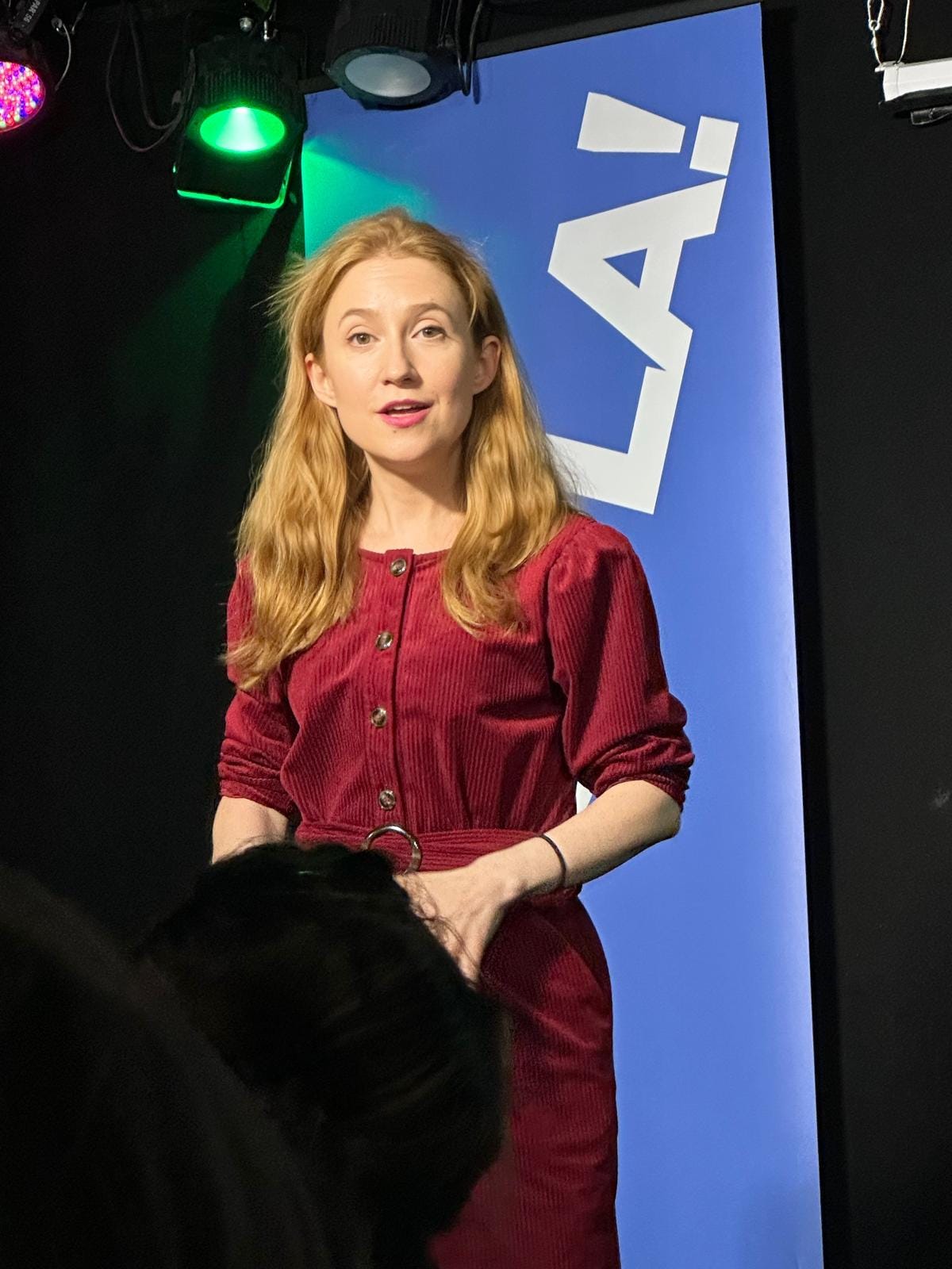a white woman with ginger hair and a red jumpsuit is on stage talking to an audience