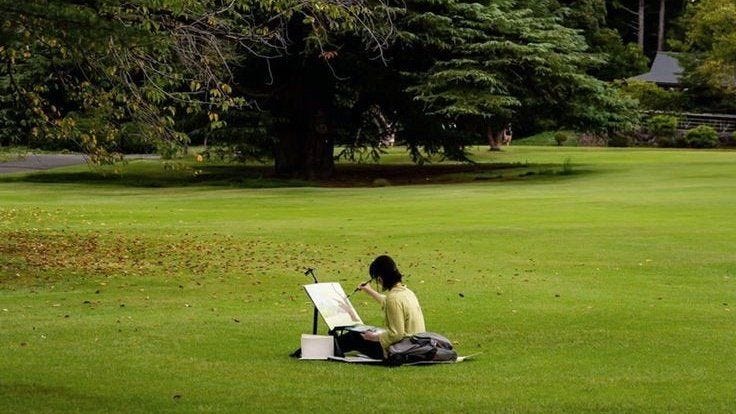 This may contain: a woman sitting on top of a lush green field next to a park filled with trees This may contain: a woman sitting on top of a lush green field next to a park filled with trees
