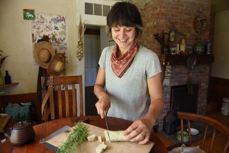 a white femme person looks down smiling at a roll of dough on a cutting board in a cozy kitchen