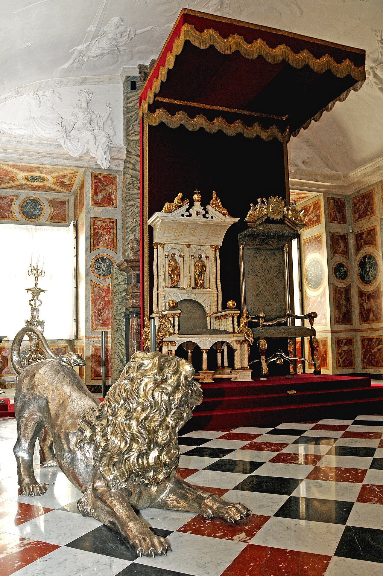 A white throne of narwhal tusk with gilt figures under a red canopy, a silver lion crouching on the checkered floor in front of it