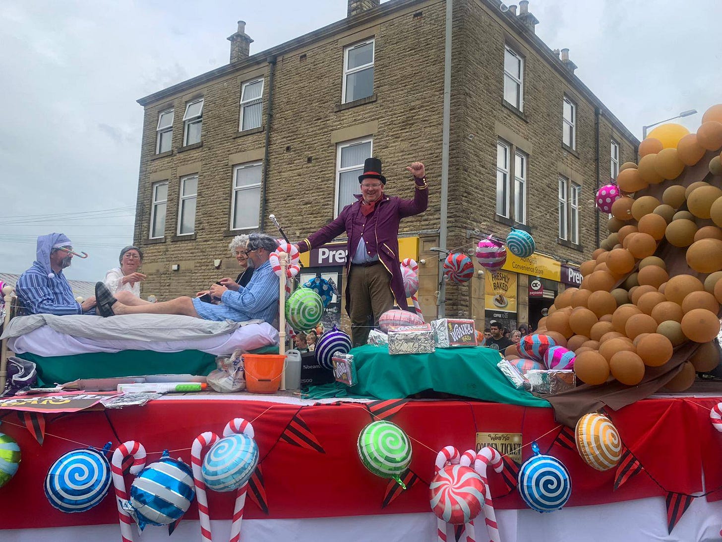 A carnival float with a man stood on the back dressed as Willy Wonka from the book "Charlie and the chocolate factory". There are also four people dressed as the grandparents from the same book. The float is decorated with balloons in the shape of sweets.