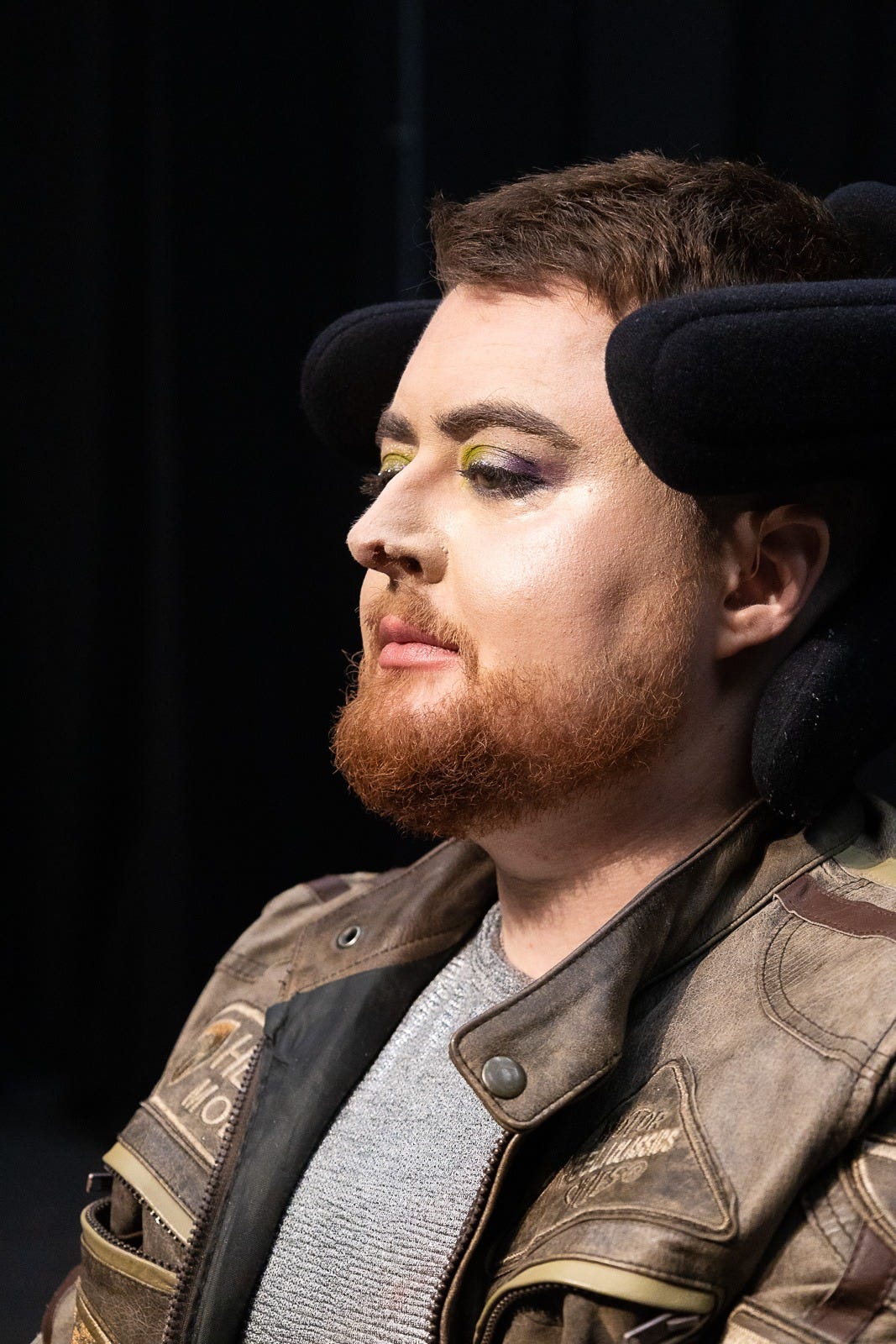 The head and shoulders of Jamie Hale, a white person in an electric wheelchair wearing green and purple eyeshadow and pale pink lipstick. They have dark red hair and a ginger beard