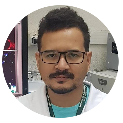 Homem com cabelos e bigode escuros, usa óculos de armação retangular e veste uma camiseta azul clara, coberta por um jaleco branco Homem com cabelos e bigode escuros, usa óculos de armação retangular e veste uma camiseta azul clara, coberta por um jaleco branco