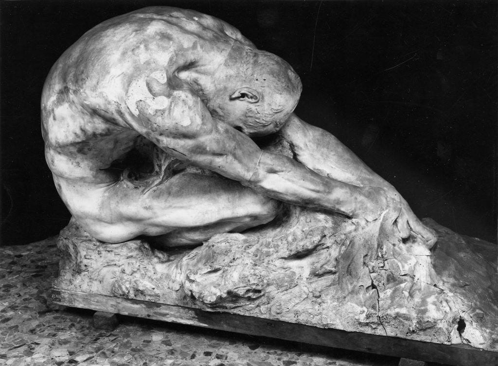 A sculpture titled "L'umanità contro il male" by Gaetano Cellini. It features a muscular, nude human figure in a powerful, strained crouch atop a rugged, rocky base. The figure is bent deeply forward, with their head lowered near the ground and powerful arms extended downward, conveying intense effort and struggle. 