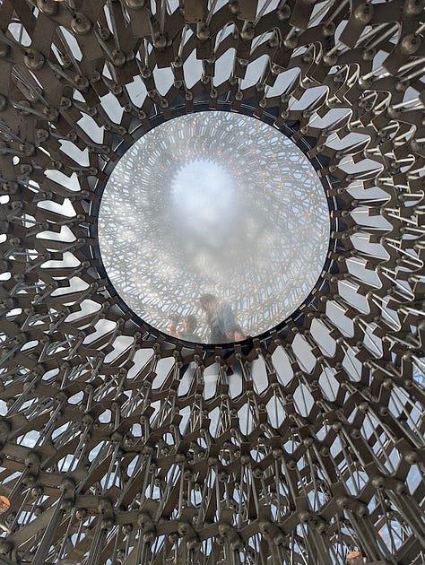 Three views of The Hive at Kew Gardens, a large aluminium lattice structure rising from a grassy landscape, shown from a distance, from beneath, and looking up through its circular interior.
