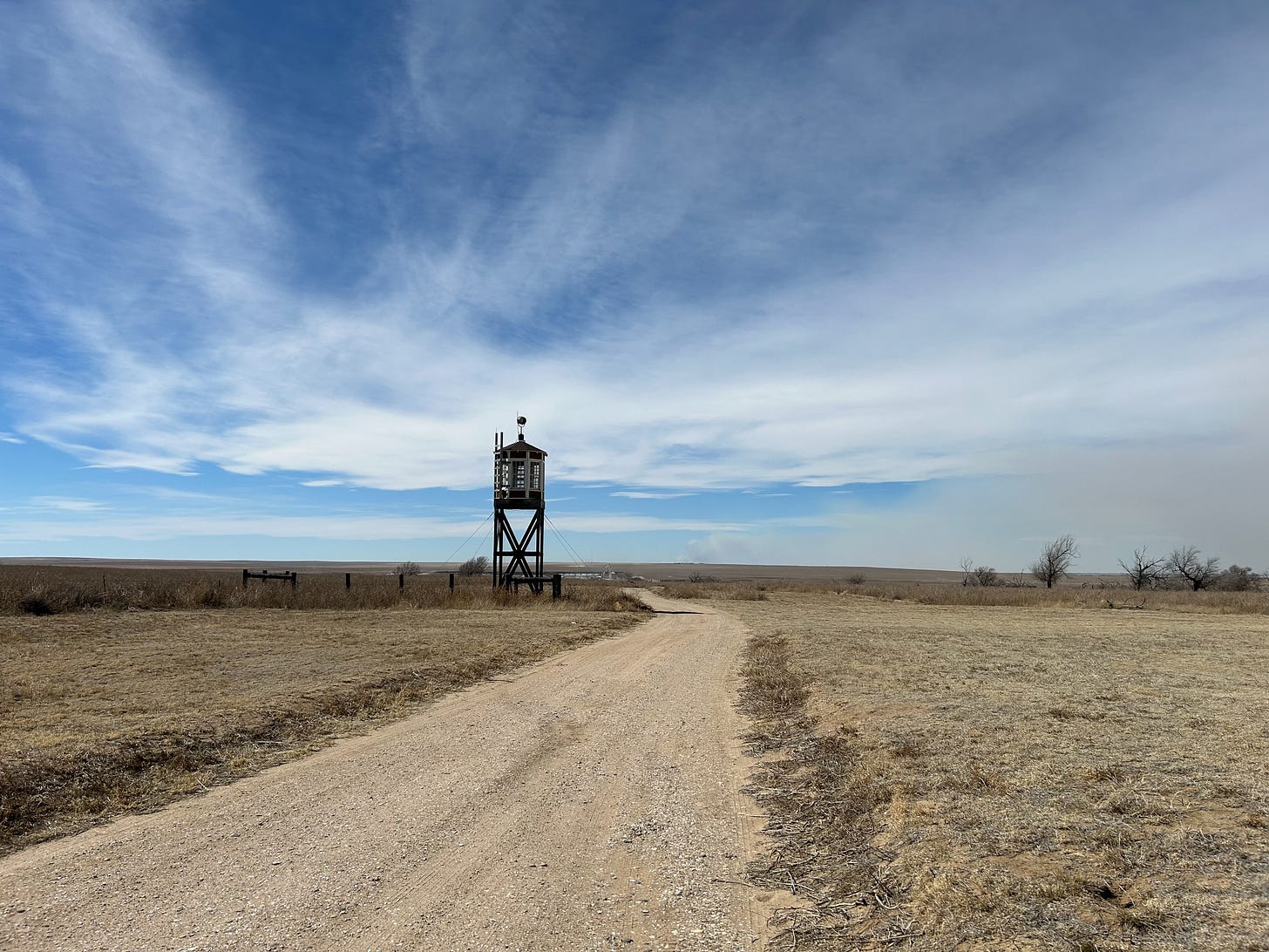 A recreation of a guard tower nearby the previous picture's residential building. The structure is wood. The base resembles the cross bracing of a coastal dock. The top, where the guard would stay reminds of European resort architecture with contrasting white and brown wooden planks. The foreground is a dirt road stretching past the guard tower in the background. The sky is a bright blue with scattered cirrus counds.