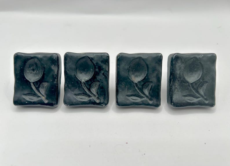 May include: Four square black ceramic tiles with a raised design of a single flower with a stem and leaves. The tiles are arranged in a row.