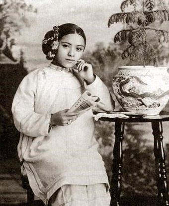Sepia-toned studio portrait of a young woman identified as Tye Leung, seated with one hand raised near her chin and the other holding an open magazine or journal, wearing a light-colored traditional jacket with flowers in her hair, beside a large patterned ceramic planter on a wooden stand, in front of a painted backdrop.