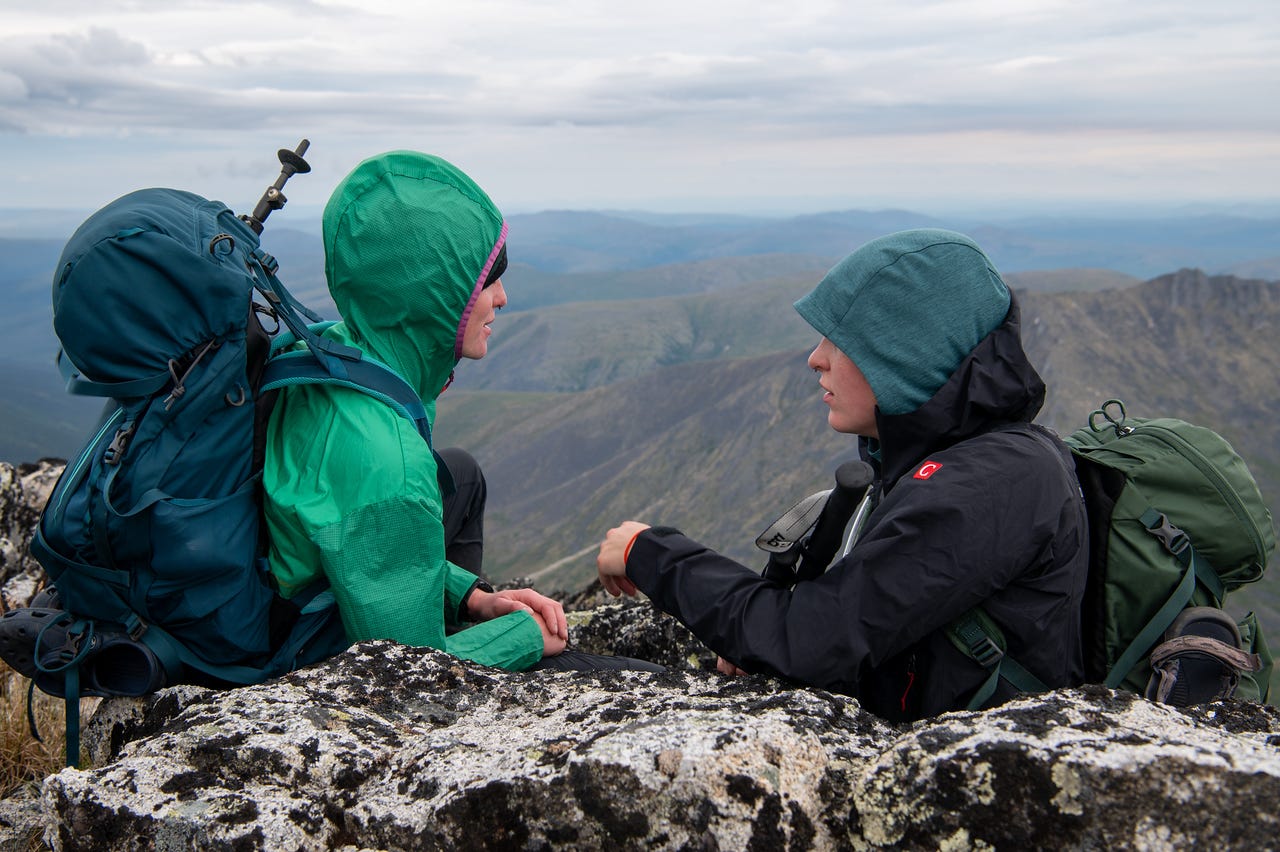 Two hikers in hooded shells sit facing each other among lichen-covered summit boulders, packs beside them, talking, with layered mountains and a wide grey sky falling away behind.
