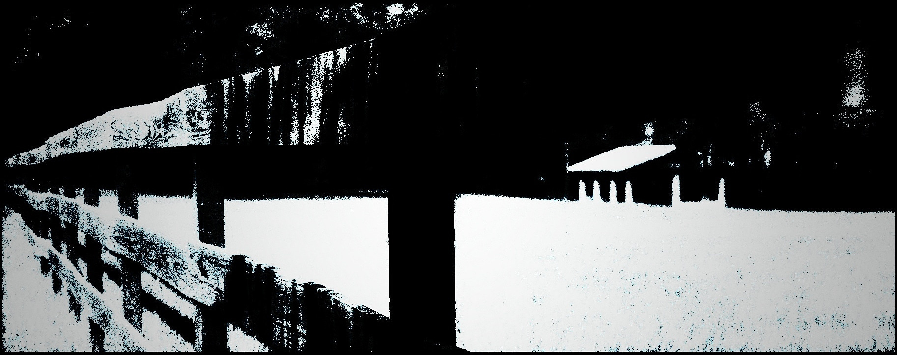 A long horizontal landscape in high-contrast black-and-white. The left half is taken up by a wooden rail fence dwindling off to a distant horizon. On the right, set against a stark white foreground, is a blurry, nondescript house, shed, garage, or pavilion; there may or may not be a large tree at the right edge of the frame. Any traces of sky are black.