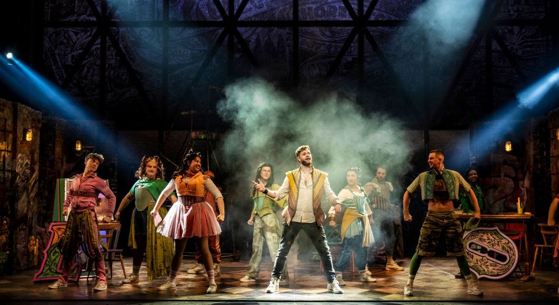 A dark blue stage with nine actors in colourful outfits, with them looking towards the centre to Jay McGuinness