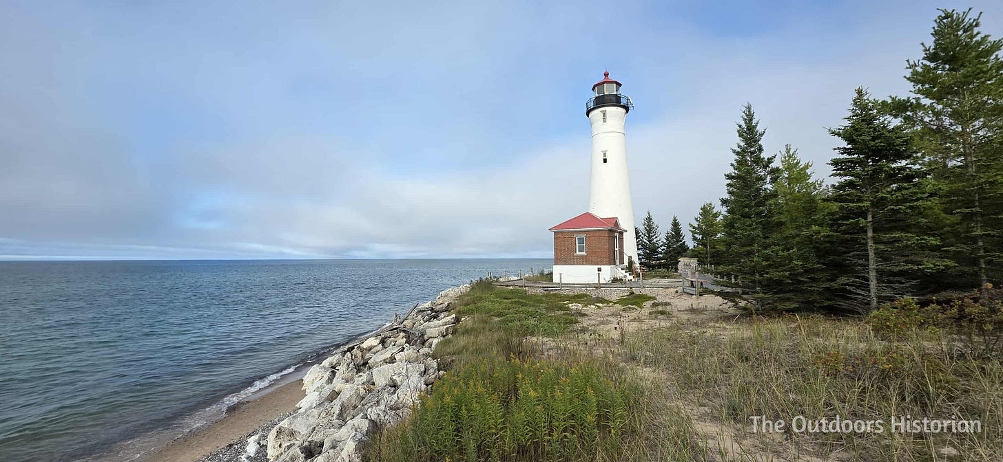 A tall, white lighthouse on a low bluff armored with riprap along a beach. There is blue sky with many white clouds in the background.