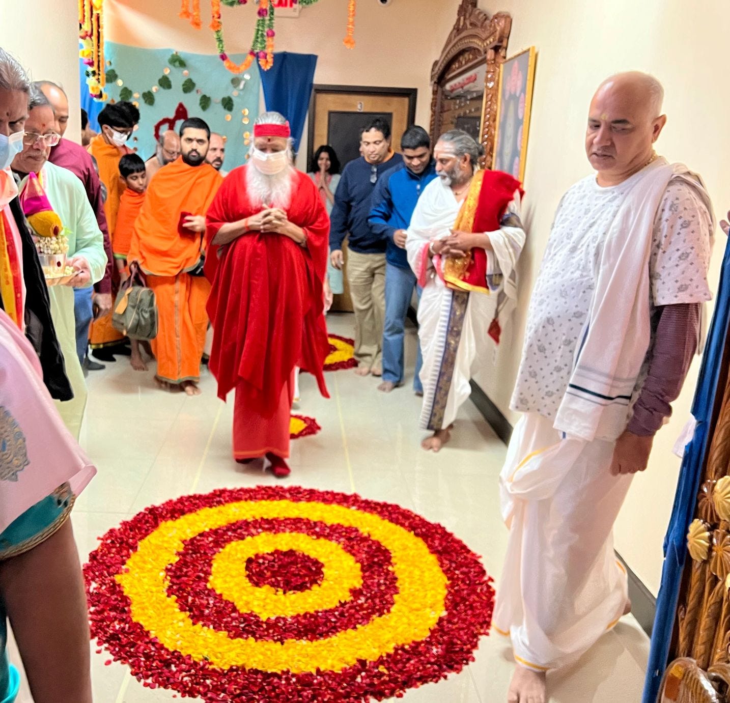 Sri Swamiji, dressed in orangey pink robes and a face mask, enters the temple with devotees behind him. Vahni is standing in the background in a light blue salwar with her hands in prayer position Sri Swamiji, dressed in orangey pink robes and a face mask, enters the temple with devotees behind him. Vahni is standing in the background in a light blue salwar with her hands in prayer position