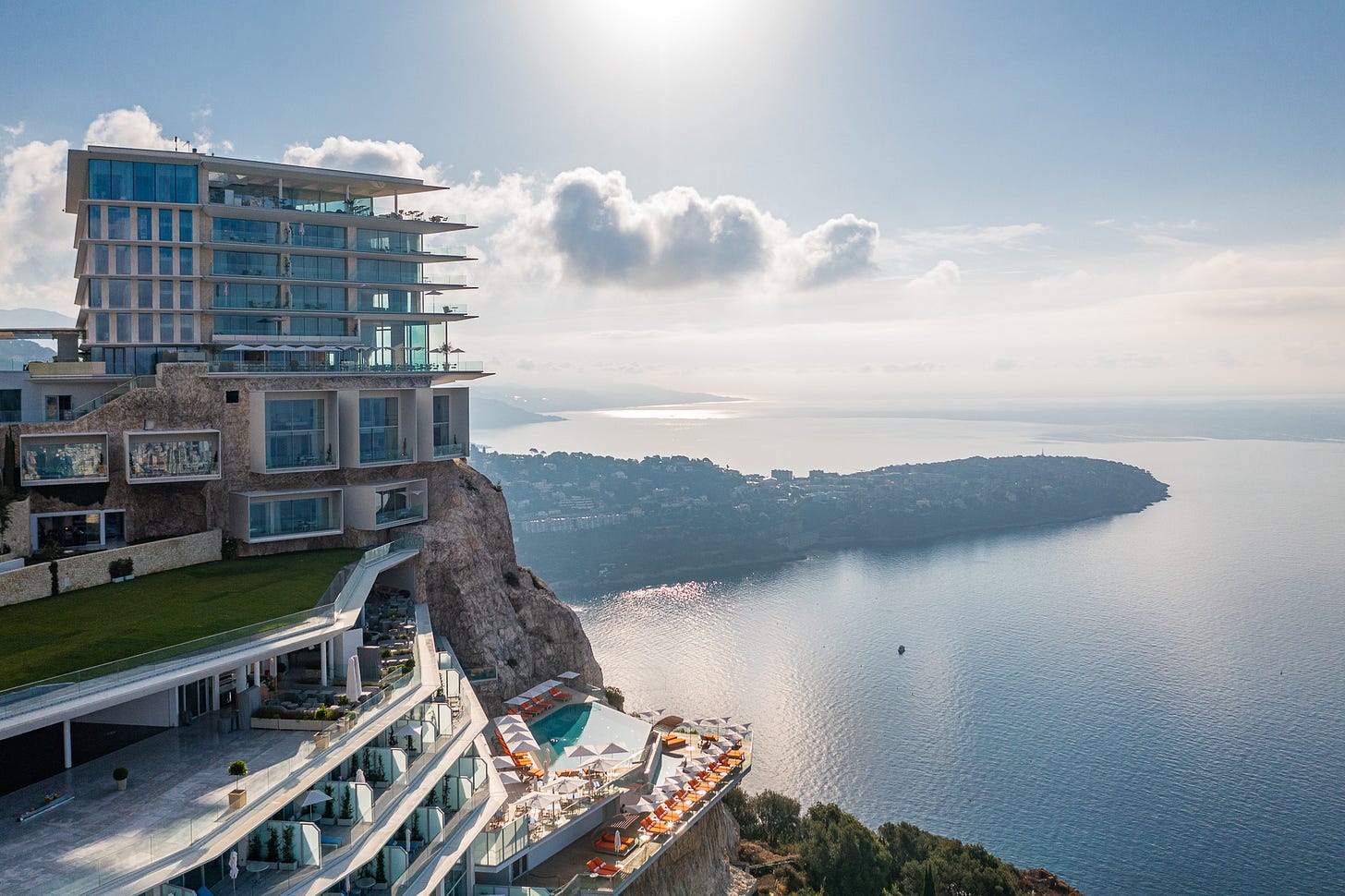 Aerial drone view of The Maybourne Riviera luxury hotel perched on a clifftop above the Mediterranean Sea on the French Riviera near Monaco. The modern multi-storey building features cascading glass balconies, a cantilevered infinity pool surrounded by orange sun loungers and parasols on a terrace overhanging the cliff edge, and a manicured green lawn. The sea stretches below toward Cap Martin and the coastline of Roquebrune-Cap-Martin under a hazy blue sky with scattered clouds and sun glinting off the water. Aerial drone view of The Maybourne Riviera luxury hotel perched on a clifftop above the Mediterranean Sea on the French Riviera near Monaco. The modern multi-storey building features cascading glass balconies, a cantilevered infinity pool surrounded by orange sun loungers and parasols on a terrace overhanging the cliff edge, and a manicured green lawn. The sea stretches below toward Cap Martin and the coastline of Roquebrune-Cap-Martin under a hazy blue sky with scattered clouds and sun glinting off the water.