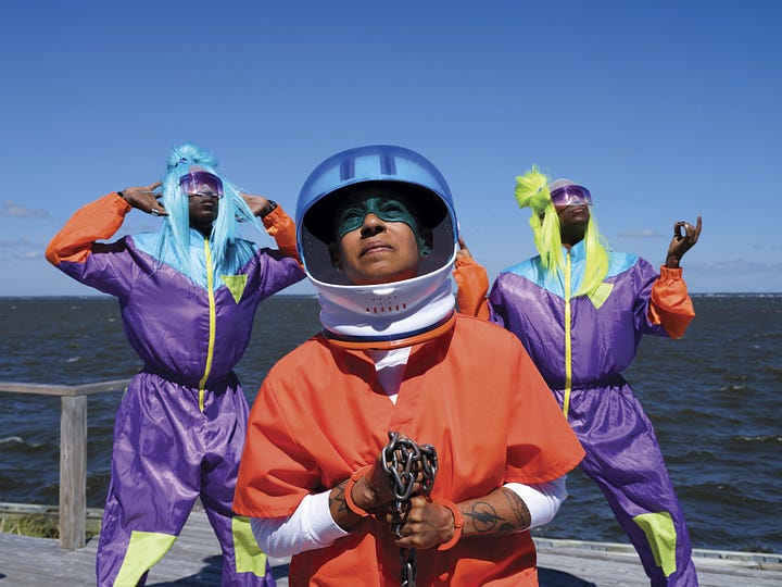 1) Three Black people in bright costumes pose by the water. The person in front. Lola, wears an orange suit, blue and white helmet, and holds chains, while two others wear colorful wigs and sunglasses. Lola is looking up at the sun and she also has orange handcuffs on their wrists. The two people in the back are wearing purple and yellow space-like suits, with blue and neon green wigs on. 2)Elderly Asian woman in a purple floral dress sits in front of her abstract black-and-white artwork. Shelves with rolled canvases are behind her, and she has a calm expression. Her hands are crossed and resting on her lap, and she is giving a slight smile to the camera. She has on long beaded necklaces, and several rings and a bracelet. She has short black hair, and she is wearing dark red lipstick.
