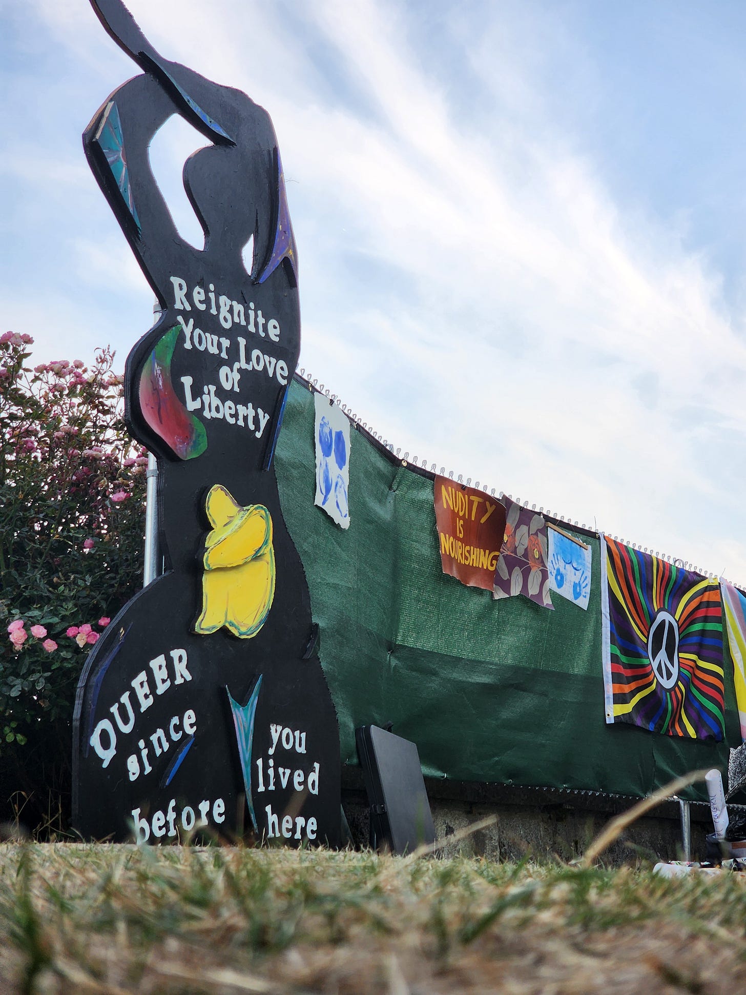 A large black painted wooden cutout of a nude figure stands at Denny Blaine Park, painted with the words "Reignite Your Love of Liberty" and "Queer since before you lived here." Behind it, green privacy tarps line a fence hung with protest signs reading "Nudity is Nourishing" and a rainbow peace flag. Pink roses bloom to the left under a partly cloudy sky. A large black painted wooden cutout of a nude figure stands at Denny Blaine Park, painted with the words "Reignite Your Love of Liberty" and "Queer since before you lived here." Behind it, green privacy tarps line a fence hung with protest signs reading "Nudity is Nourishing" and a rainbow peace flag. Pink roses bloom to the left under a partly cloudy sky.