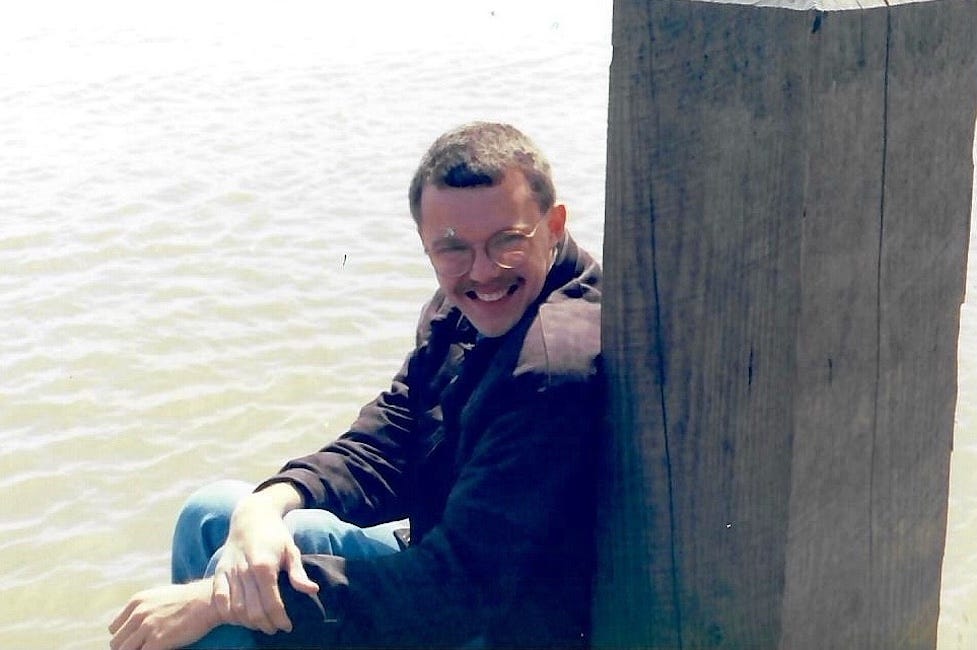 A young man sits by a wooden post at the edge of a body of water, smiling in bright sunlight. He wears round glasses, a dark jacket, and jeans, with his hands loosely clasped around his knees. The water behind him is calm and softly rippled. A young man sits by a wooden post at the edge of a body of water, smiling in bright sunlight. He wears round glasses, a dark jacket, and jeans, with his hands loosely clasped around his knees. The water behind him is calm and softly rippled.