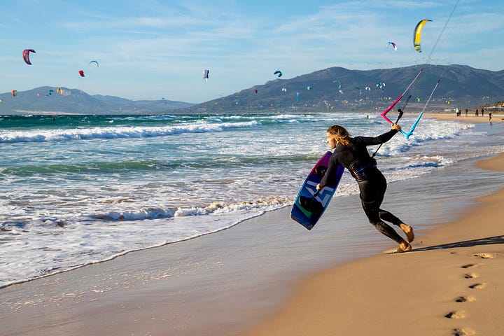Kitesurfer riding near the medieval castle walls in Tarifa; the Mar Mediterráneo and Océano Atlántico signs marking where two seas meet at the Tarifa causeway; a kitesurfer suspended mid-air above the Strait of Gibraltar; a man running into the surf with a kiteboard on Tarifa beach