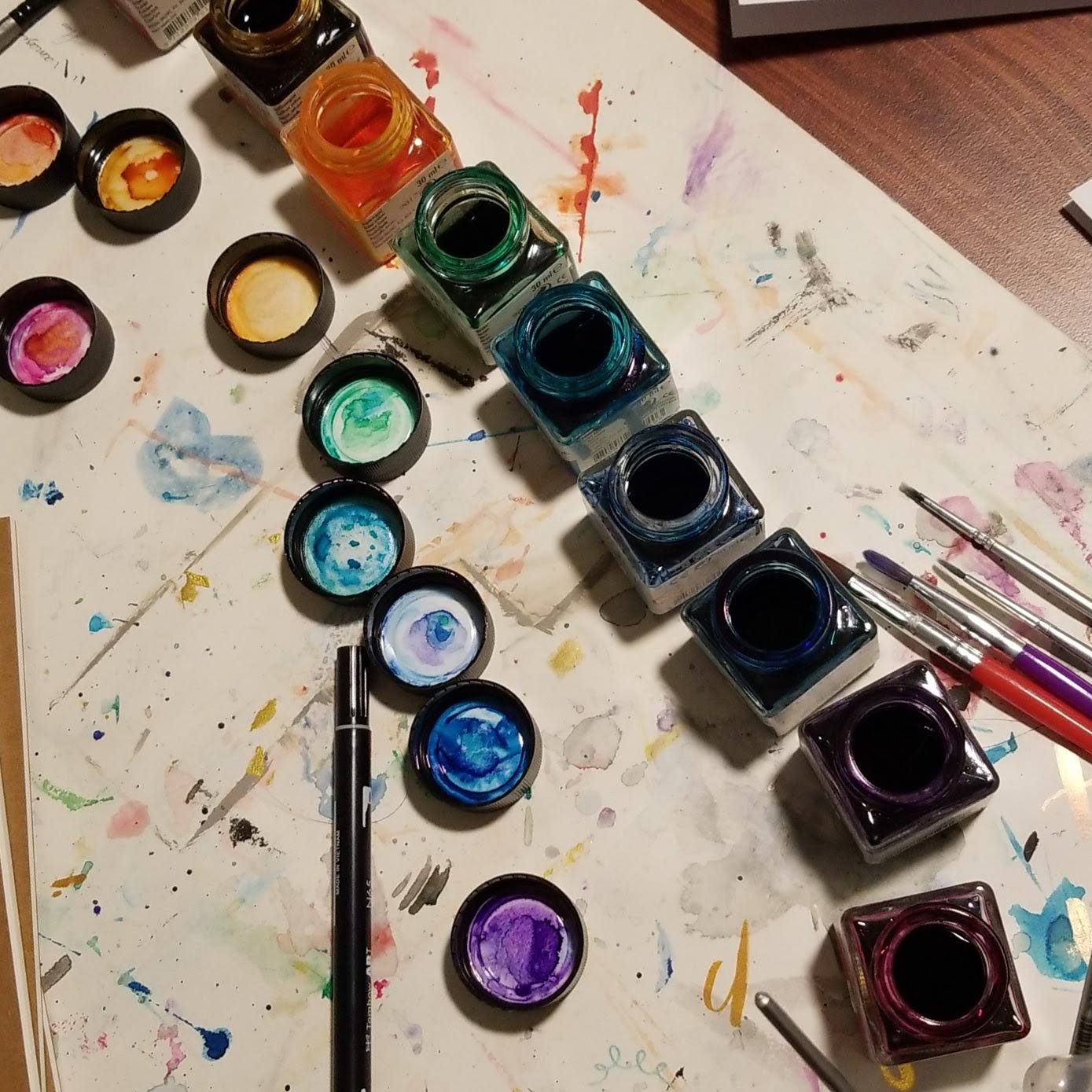 Overhead view of an artist's workspace with a row of open glass ink bottles in various vibrant colors including red, orange, green, blue, and purple. Each bottle has its corresponding cap laid out in a diagonal line, displaying ink-stained lids with watercolor-like stains. The white surface beneath is heavily marked with colorful splatters and brush strokes. A black marker and several paintbrushes with colorful handles lie scattered on the table. The setting is informal and creative, suggesting an ongoing art project.
