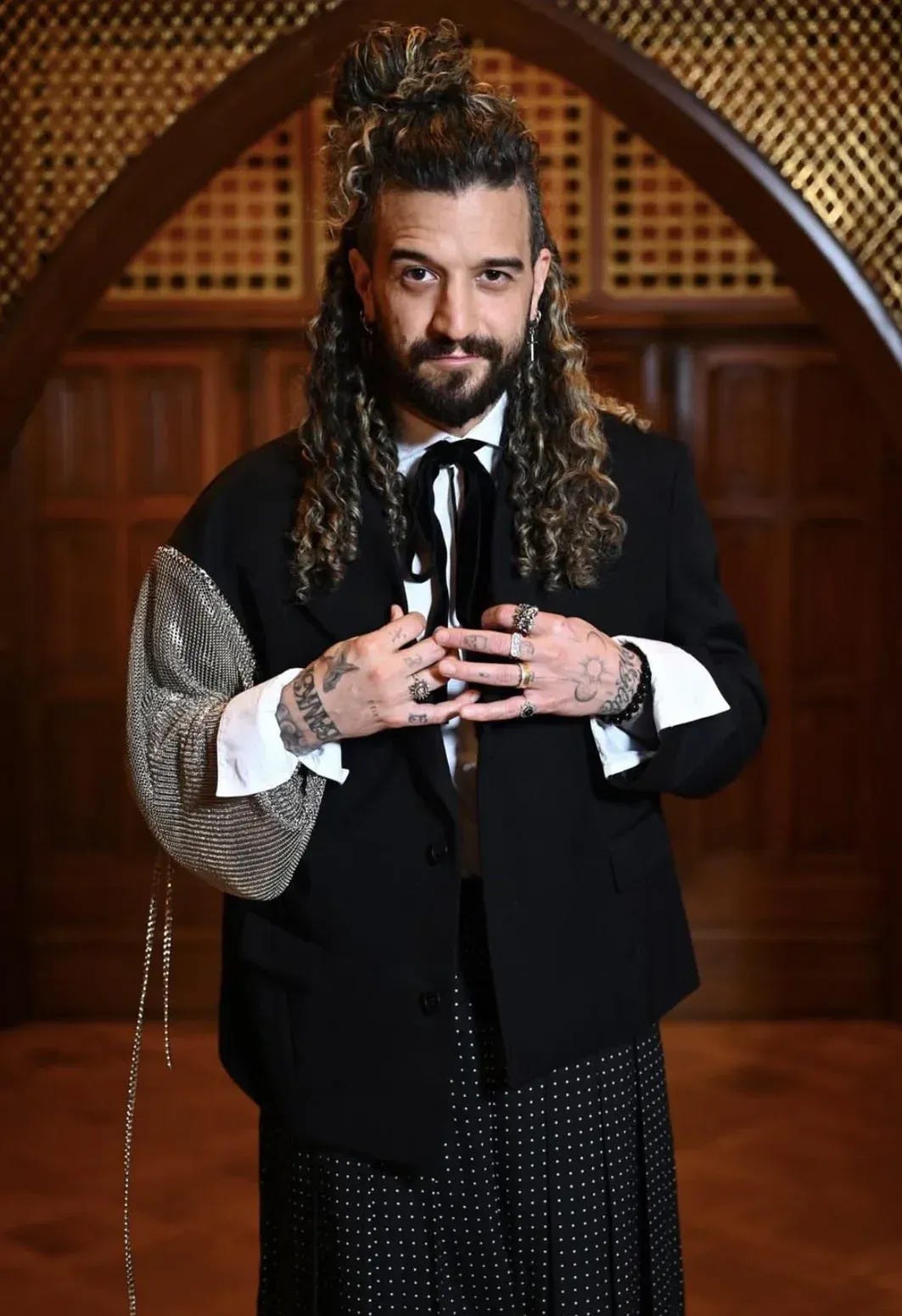 Mark in an asymmetrical black blazer with one metal mesh sleeve, black pussy-bow tie, black studded skirt, curly hair half-up, multiple rings.